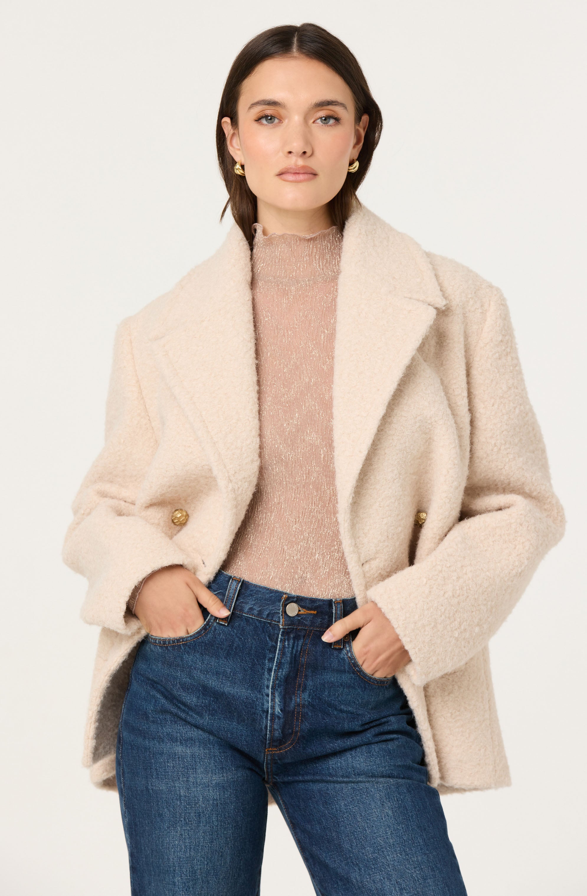 A woman is wearing a light beige, textured, oversized wool coat with wide lapels and gold button details on the sleeves. She has on a pink, shimmery, high-neck top underneath. She is also wearing high-waisted blue jeans with a classic cut.