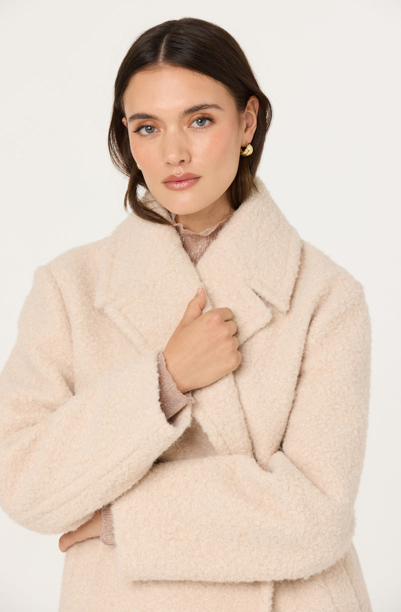 A woman wearing a soft, cream-colored, textured coat with a wide collar. The coat appears warm and cozy. She has small, gold hoop earrings and is holding the lapel of her coat with her right hand.