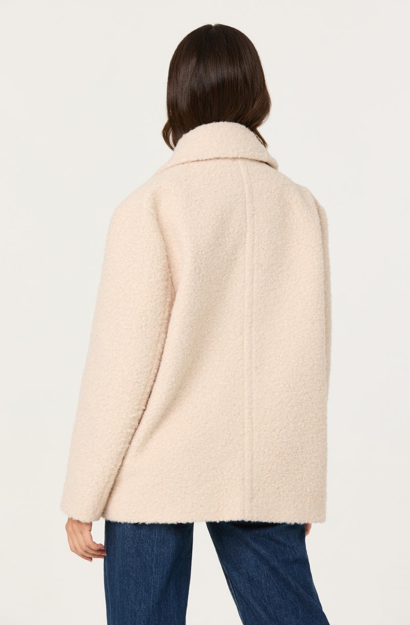 A woman is wearing a cream-colored, textured, wool-like coat with a wide collar. The coat appears soft and plush, with a straight cut that extends to the hips. She is also wearing dark blue jeans.