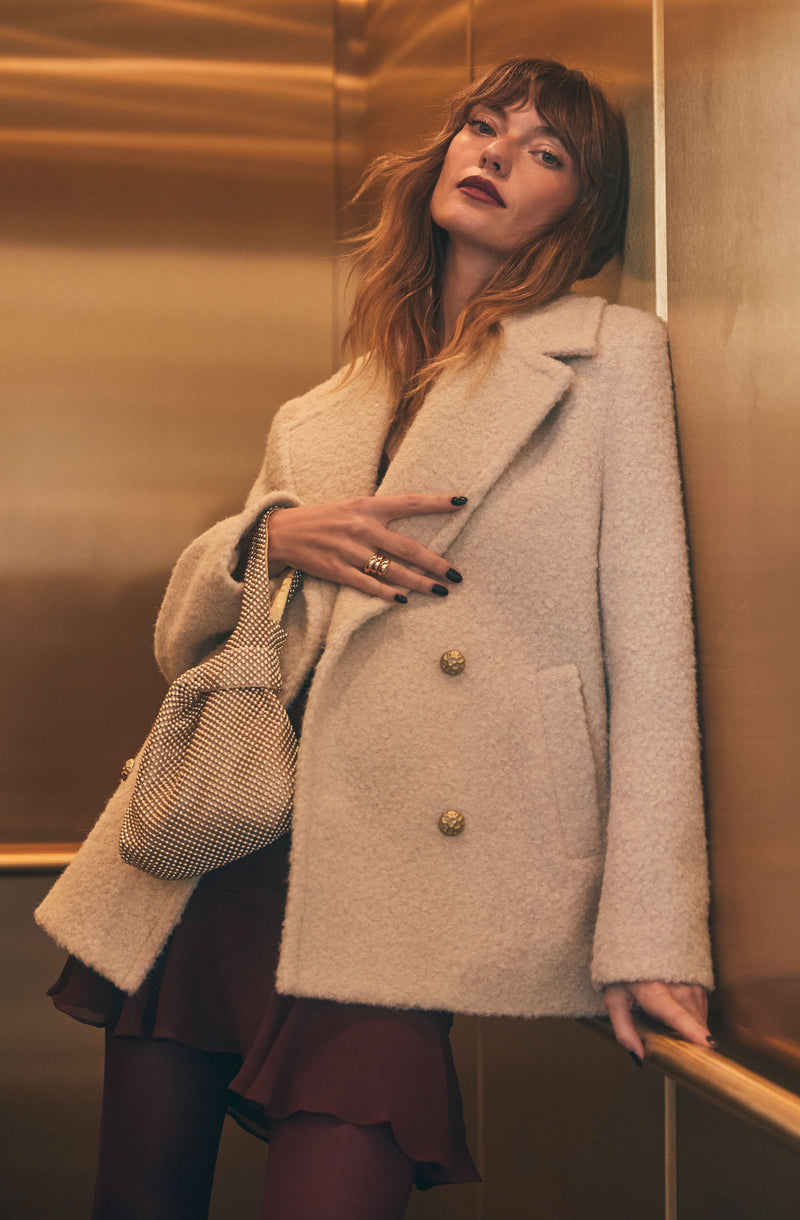 A woman is wearing a beige, double-breasted, wool coat with large gold buttons. She has on dark lipstick and a subtle makeup look, and holds a small, patterned handbag. She is also wearing a dark-colored dress with a ruffled hem and dark tights.