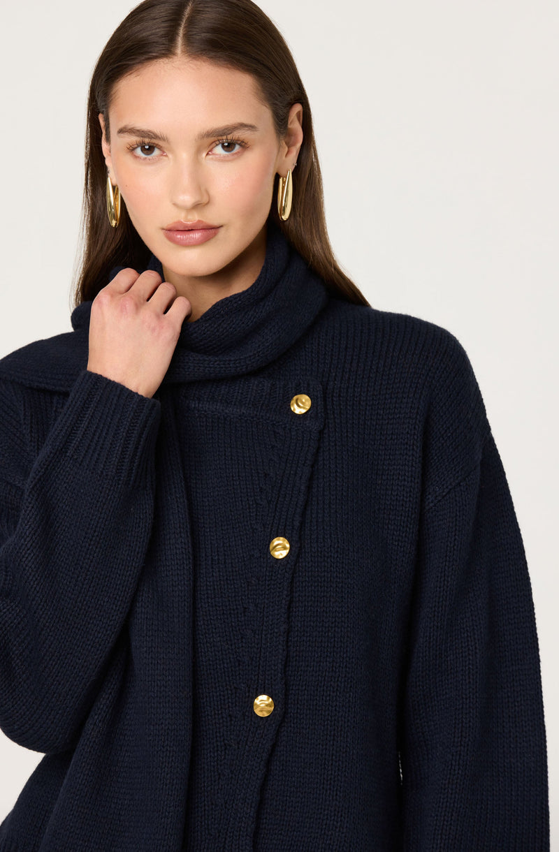 A woman is wearing a dark navy blue knit sweater with a turtleneck collar. The sweater features gold buttons down the front and has a textured, chunky knit pattern. She is also wearing large gold hoop earrings.