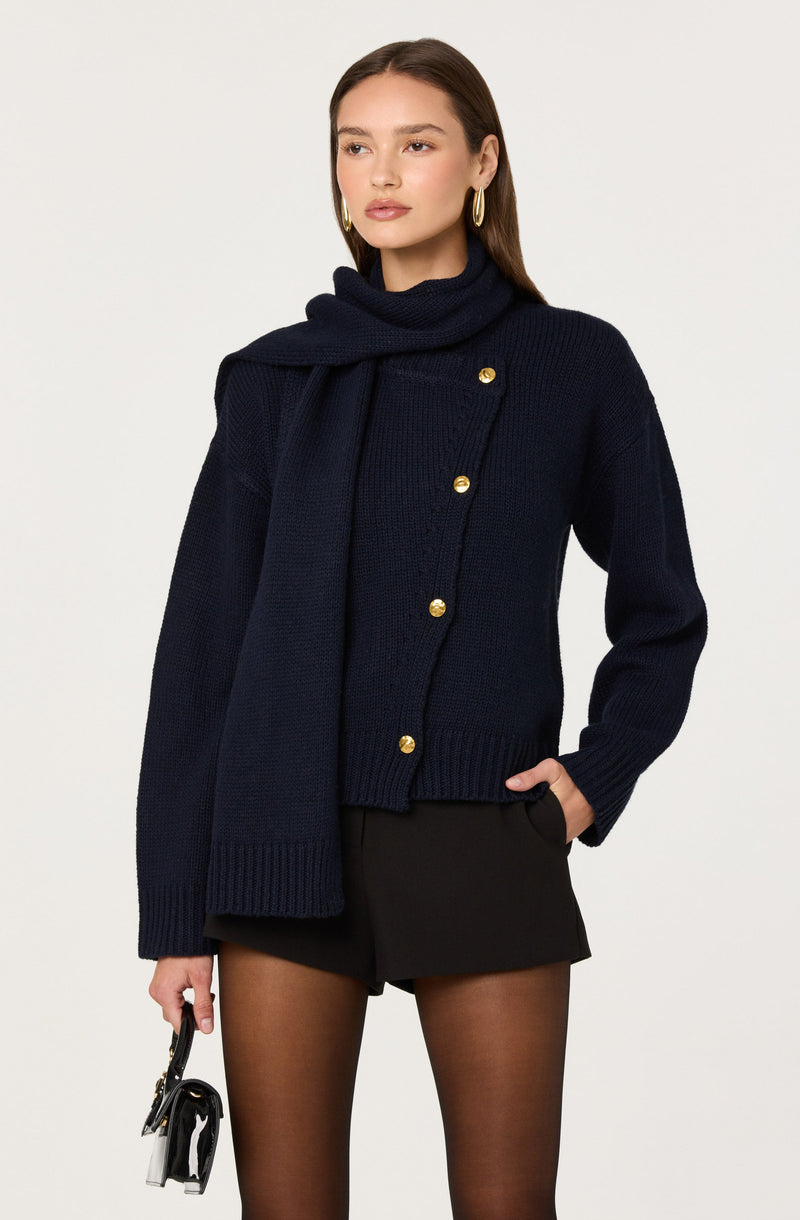 A woman is wearing a navy blue, chunky knit cardigan with large gold buttons down the front. She has a matching navy blue scarf draped over her shoulders. She is also wearing black shorts and sheer black tights. She accessorizes with large gold hoop earrings and holds a small black handbag in her left hand.