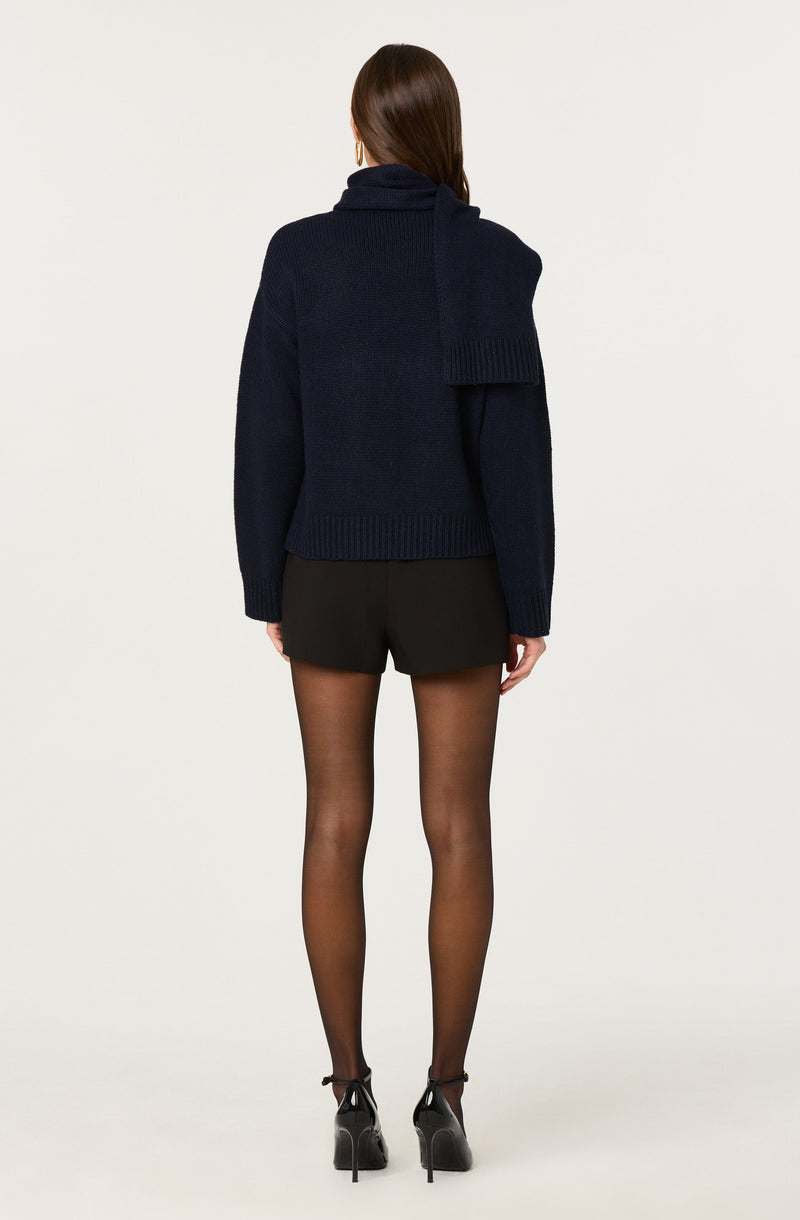 A person is wearing a dark navy, oversized turtleneck sweater with a large scarf detail draped over one shoulder, paired with black shorts, sheer black tights, and black high-heeled shoes with ankle straps.