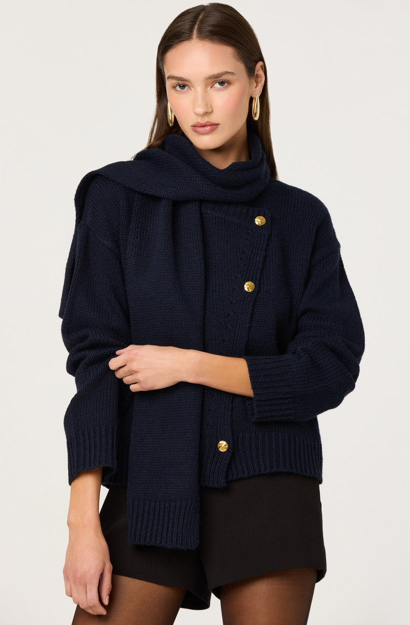 A woman is wearing a navy blue knit sweater with gold buttons down the front. She has a matching navy blue scarf draped over her shoulders and tied loosely around her neck. She is also wearing large gold hoop earrings. The sweater has a chunky knit texture with ribbed cuffs and hem.