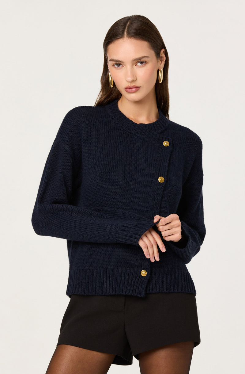 A woman wearing a dark navy blue knitted cardigan with a round neckline and gold buttons down the front. The cardigan has long sleeves and a ribbed hem. She is also wearing black shorts and gold hoop earrings.