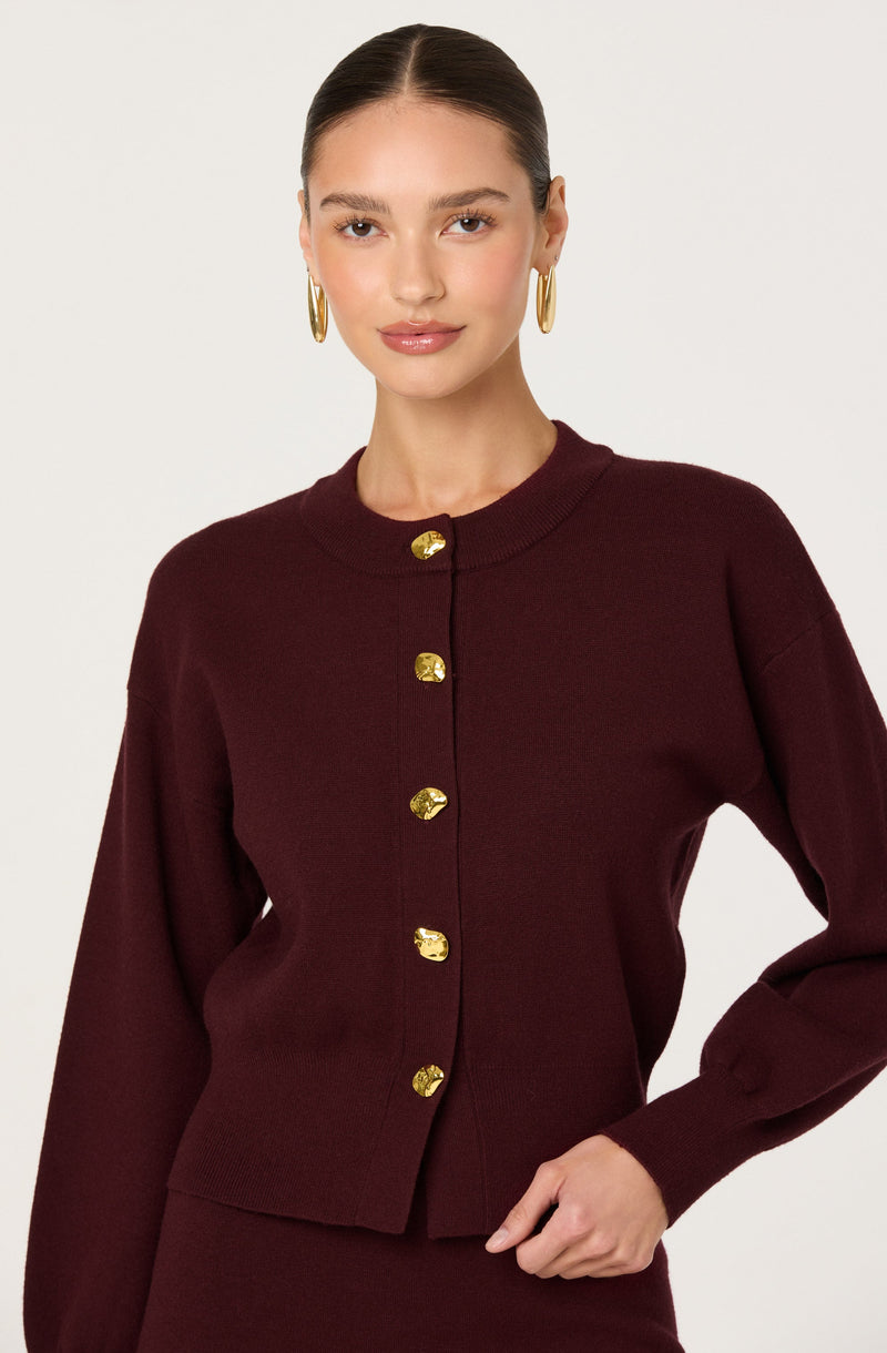 A woman is wearing a dark burgundy cardigan with gold buttons down the front. The cardigan has a round neckline, long sleeves, and a ribbed hem. She is accessorized with large gold hoop earrings and has her dark hair pulled back in a sleek style.