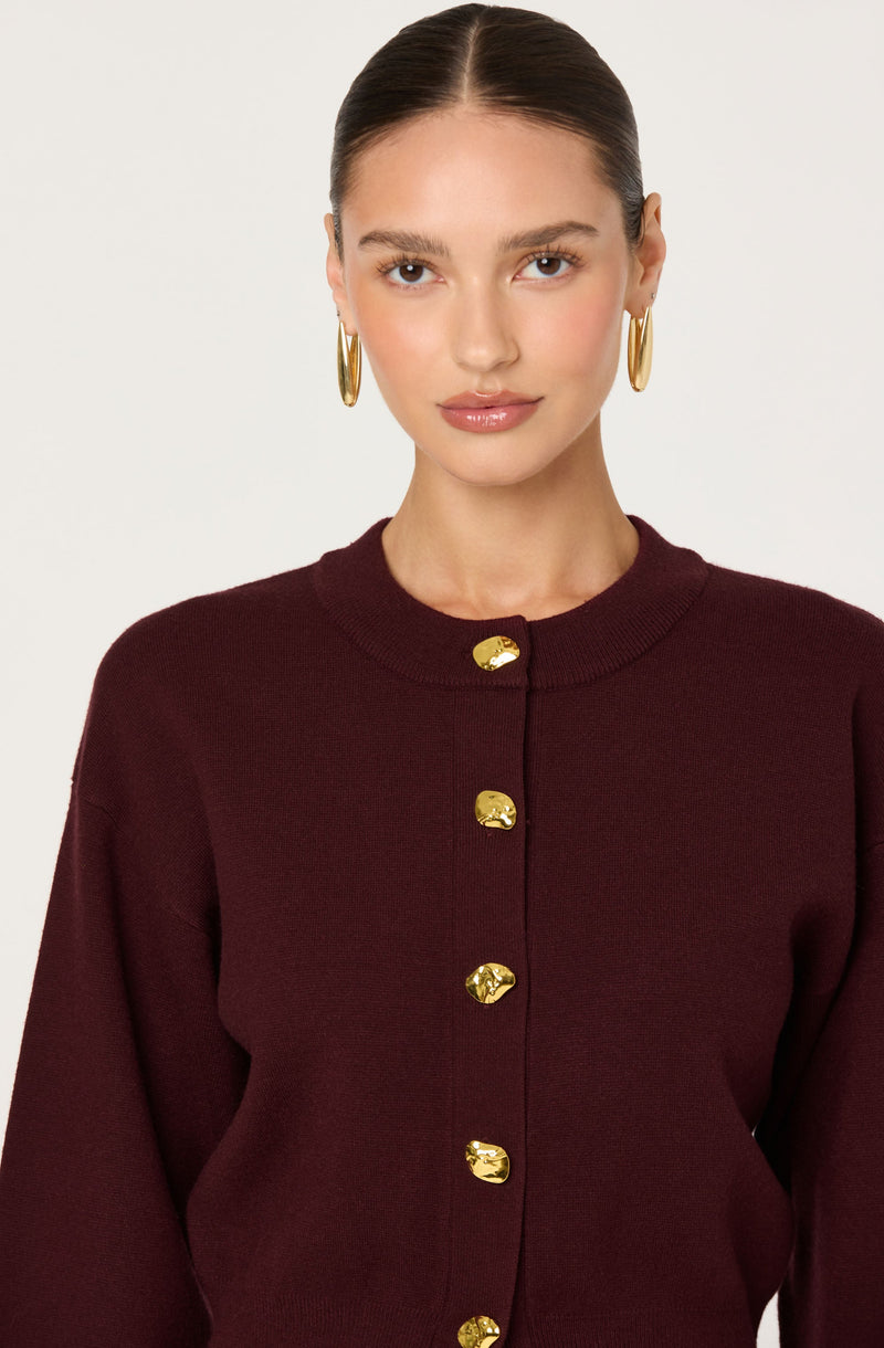 A woman is wearing a dark burgundy cardigan with gold buttons down the front. She has gold hoop earrings and her hair is pulled back in a sleek style.