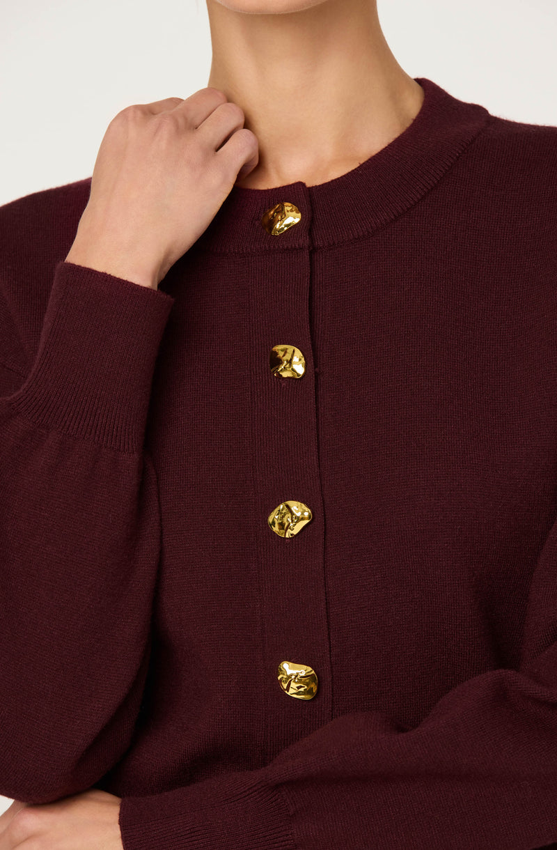 A person is wearing a dark red or burgundy knit cardigan with a round neckline. The cardigan features a vertical row of shiny, gold-colored buttons down the front. The long sleeves are cuffed at the wrists.