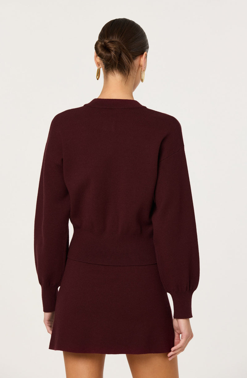 A woman is wearing a dark burgundy sweater with long, slightly puffed sleeves that are gathered at the cuffs. The sweater has a round neckline and a fitted waist. She is also wearing a matching burgundy skirt that is short in length, ending above the knees. The outfit is complemented by gold hoop earrings.