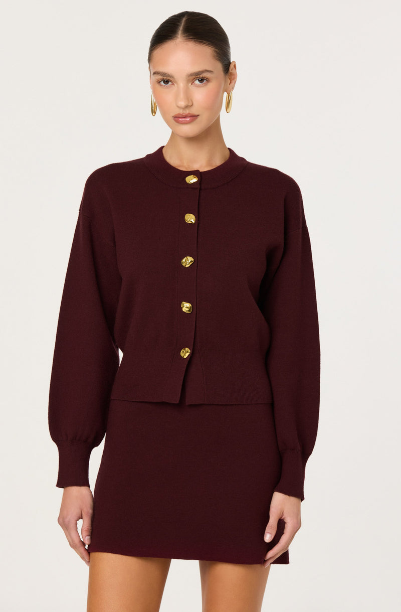 A woman is wearing a burgundy knit outfit consisting of a long-sleeve, button-up top with a round neckline and gold buttons down the front. The top has slightly puffed sleeves gathered at the cuffs. She is also wearing a matching fitted mini skirt.