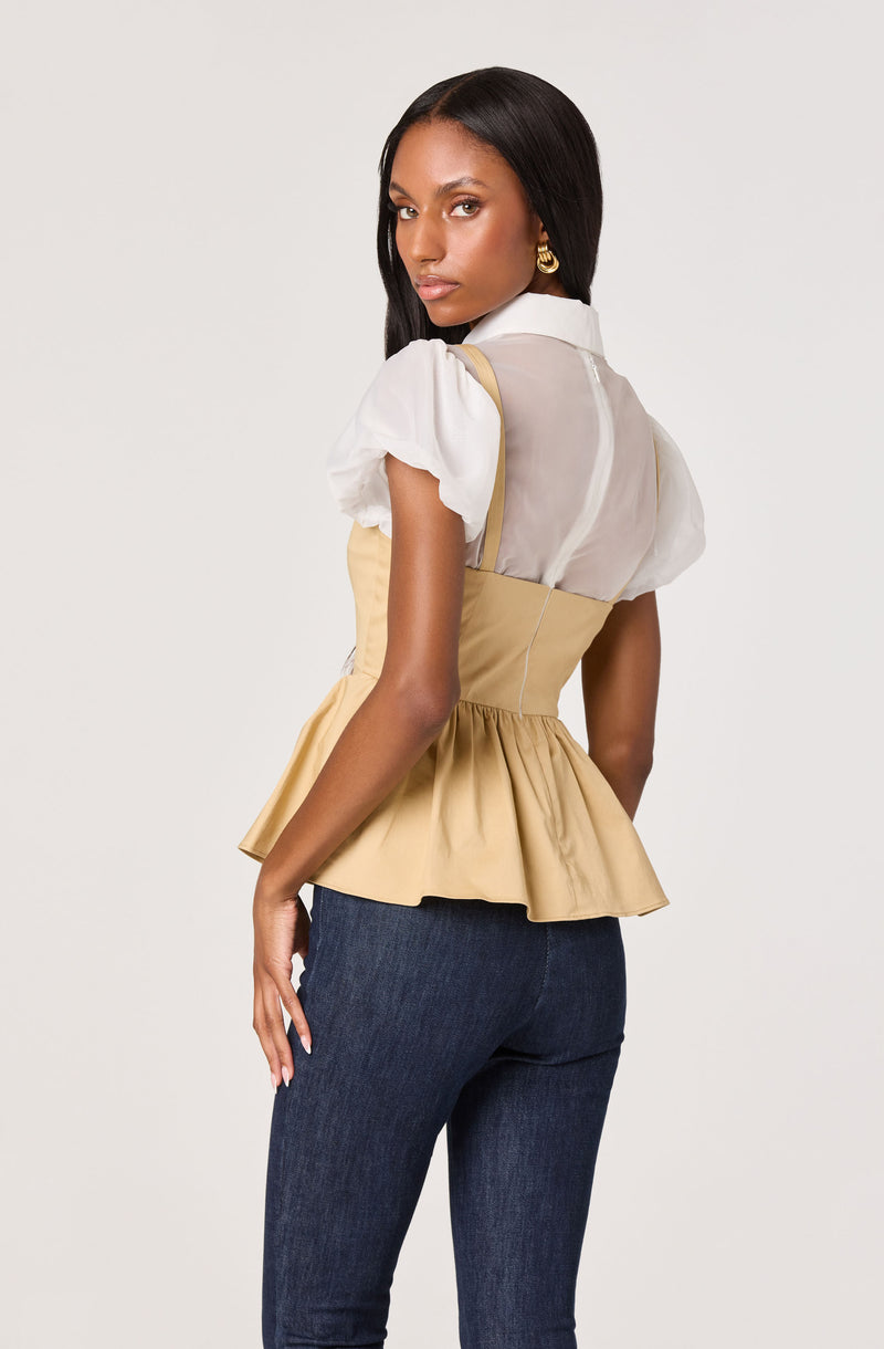 A woman is wearing a beige peplum top with a fitted waist and a flared hem. The top has a white collar and sheer white puff sleeves. She is also wearing dark blue jeans.