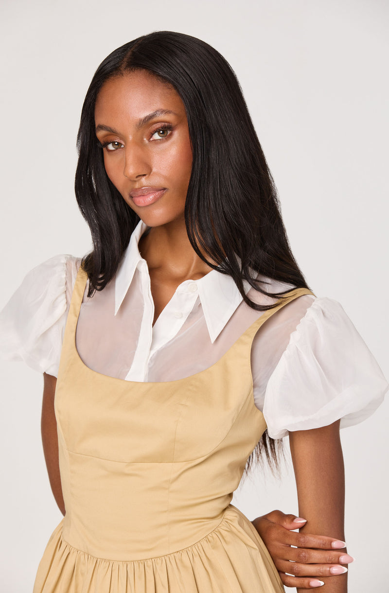 A woman is wearing a white collared shirt with sheer, puffy short sleeves underneath a beige, sleeveless dress with a fitted bodice and a gathered skirt.