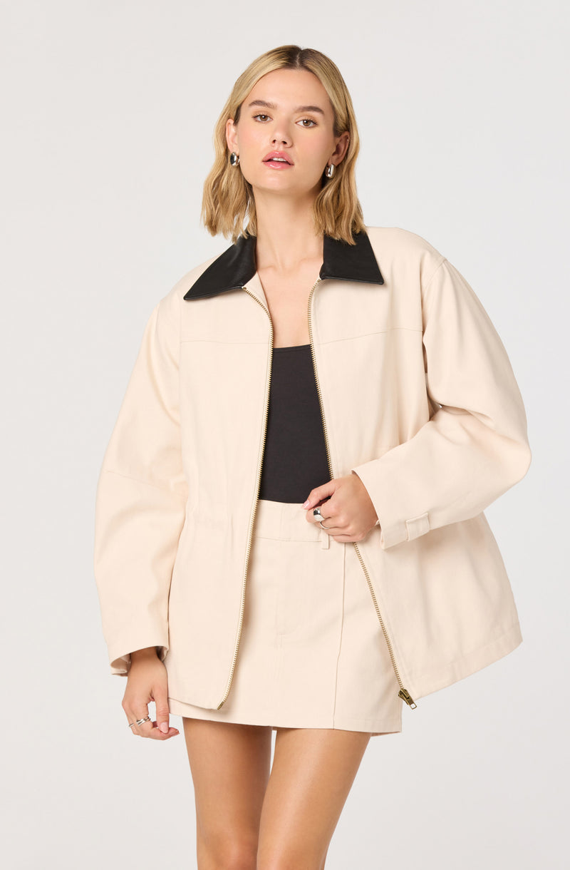 A woman is wearing a beige oversized jacket with long sleeves and a large black collar, over a black top. She has on a matching beige mini skirt with a front zipper, and silver hoop earrings.