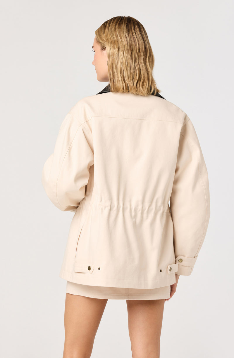 A woman is wearing a cream-colored, oversized jacket with a black collar. The jacket features a gathered waist and snap buttons at the cuffs and hem. She is also wearing a matching cream skirt underneath.