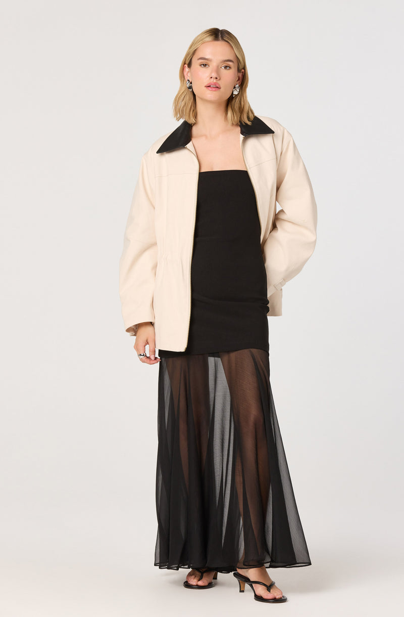 A woman is wearing a cream-colored jacket with a black collar over a fitted black strapless dress. She is also wearing sheer black wide-leg pants, which reveal her legs and black high-heeled sandals.