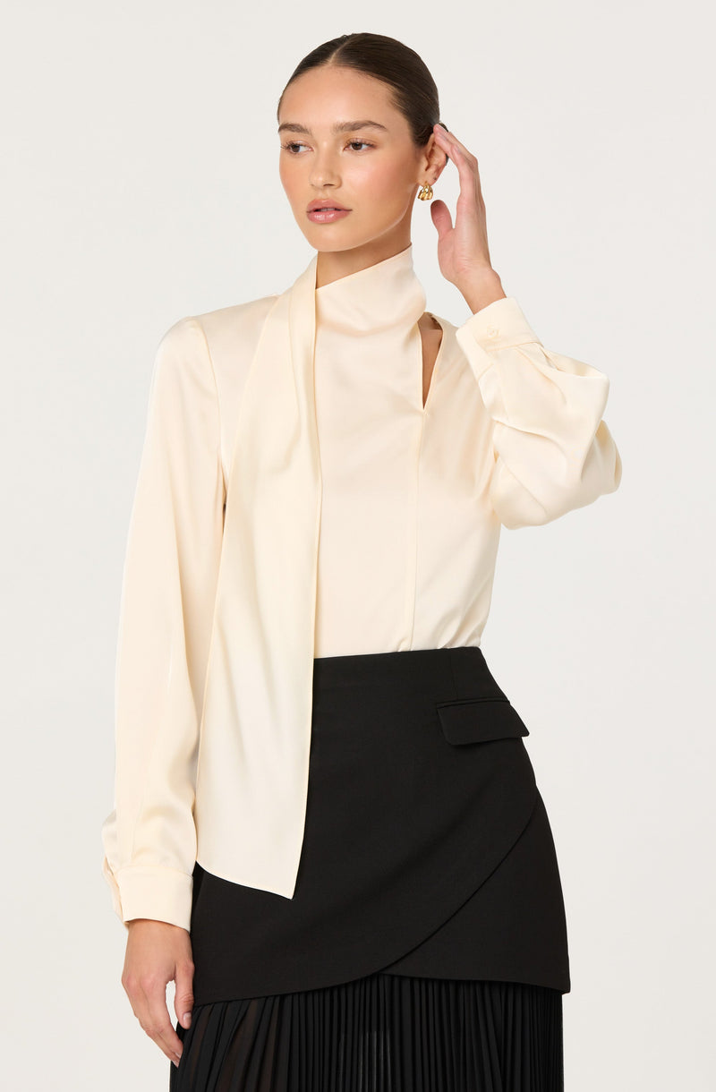 A woman is wearing a cream-colored, long-sleeved blouse with a high collar and a matching scarf-like element draped over one shoulder. She pairs it with a high-waisted, black skirt that features an asymmetrical wrap design, a flap pocket on the side, and a pleated section at the bottom. The outfit appears elegant and sophisticated.