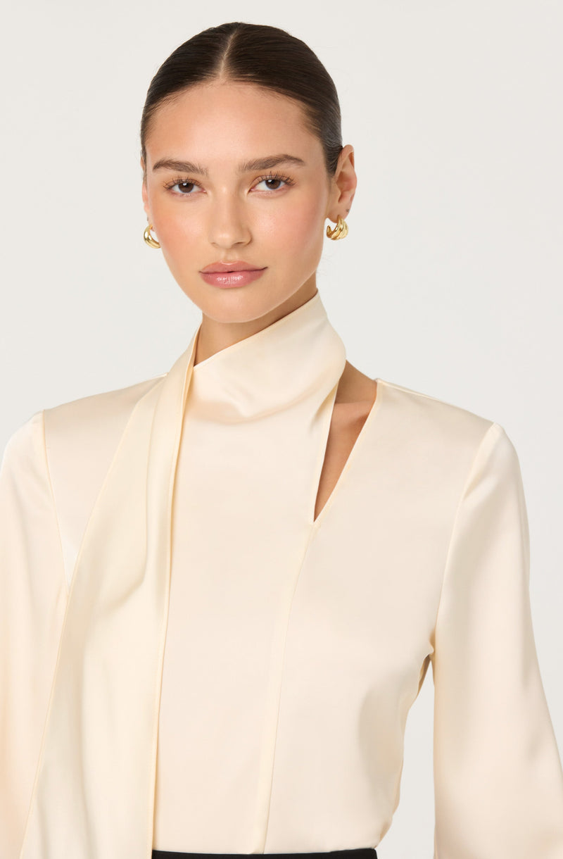 A woman is wearing a cream-colored, silky blouse with a high neckline that features a tie detail and an asymmetrical cutout on the shoulder. She has dark hair pulled back and is accessorized with gold hoop earrings.