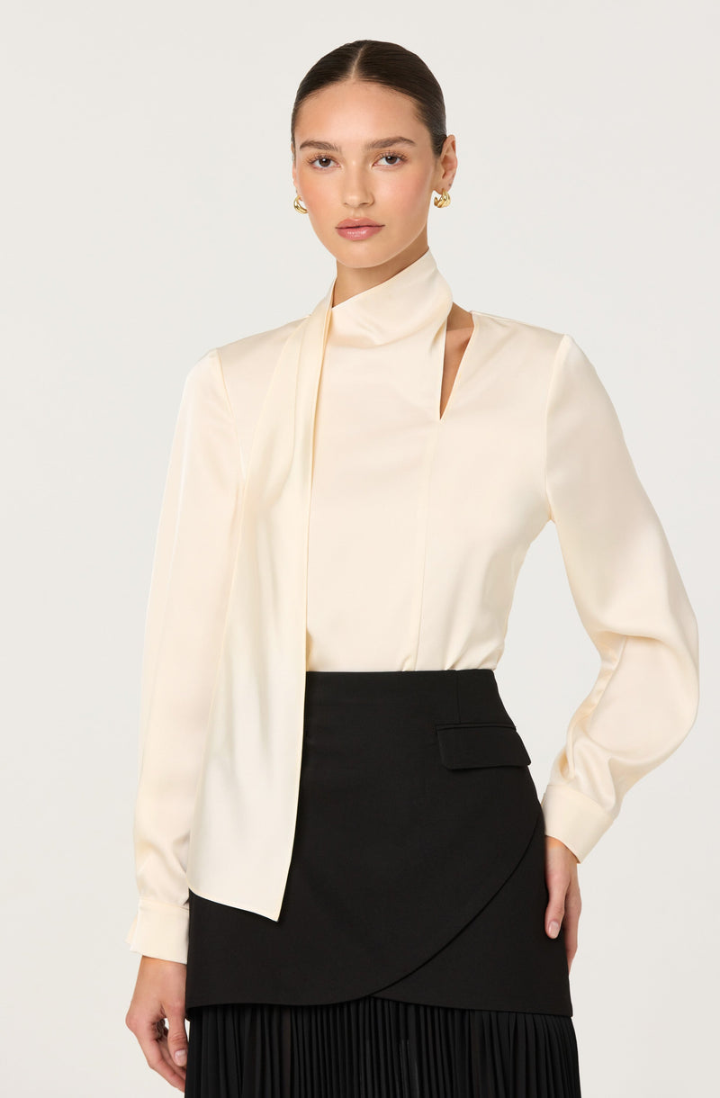 A woman is wearing a long-sleeved, cream-colored satin blouse with a high neck and a draped detail on the left side. The blouse has a smooth, shiny texture and a clean, elegant design. She pairs it with a high-waisted black skirt featuring an asymmetrical wrap style, a large front pocket, and a pleated hem.