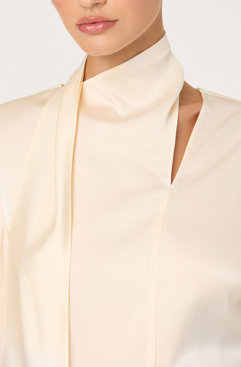 A woman is wearing a high-neck, long-sleeved cream-colored satin blouse with a slight sheen. The blouse features a small slit on the shoulder and a subtle vertical seam detail.