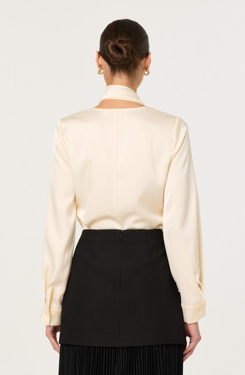 A woman is wearing a long-sleeved, cream-colored satin blouse with a high neckline and a slit at the back for closure. The blouse has a smooth, shiny texture. She is also wearing a high-waisted, black skirt that has a fitted waist and extends below the hips, with a pleated hem at the bottom.