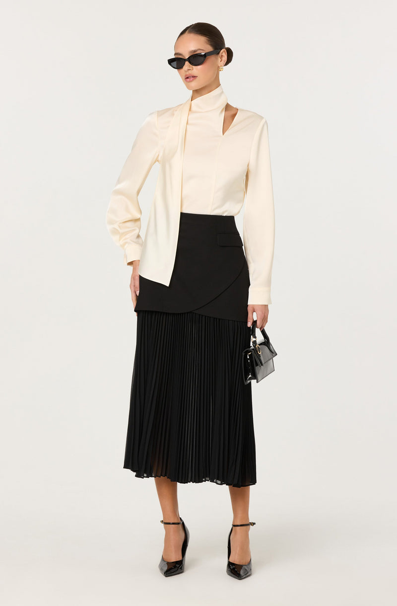 A woman is wearing a cream-colored, long-sleeve blouse with a high neckline and cut-out details on the shoulders. She has paired it with a black, asymmetrical, layered skirt that has a slight texture and pleats. She is also wearing black high-heeled shoes with ankle straps and is holding a small, structured black handbag. She is accessorized with black sunglasses and gold earrings, and her hair is slicked back.