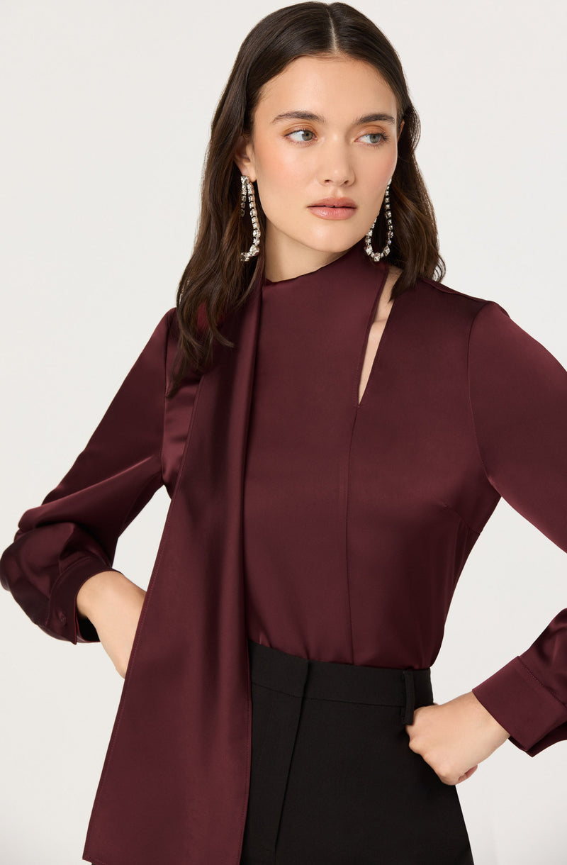 A woman is wearing a burgundy satin blouse with a high neckline and a subtle slit design near the collarbone, paired with black high-waisted pants. She has on large, dangling earrings that appear to be made of clear crystals or rhinestones.