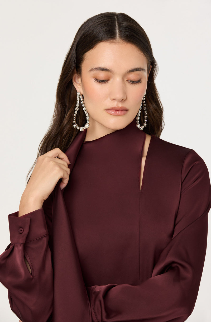 A woman with long dark hair wearing a deep burgundy satin dress with long sleeves, a small slit near the neckline, and a subtle seam running down the front. She is accessorized with large, sparkling earring hoops.