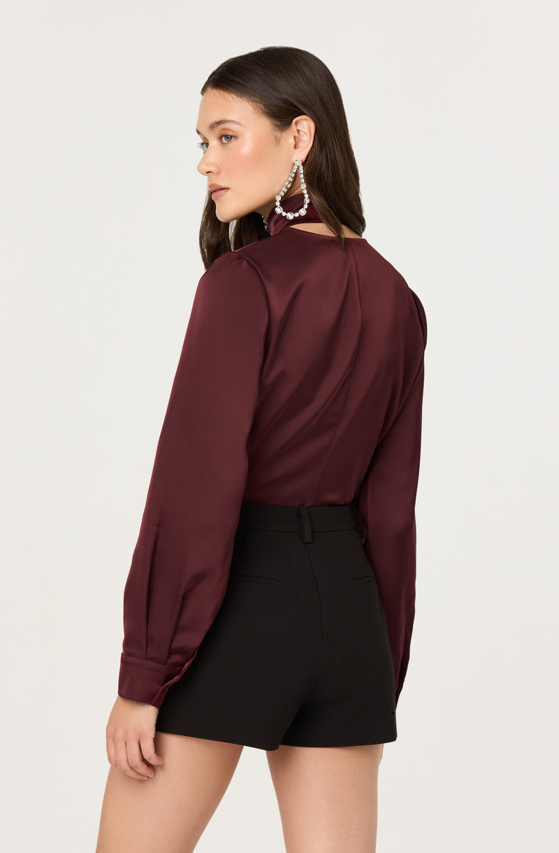 A woman is wearing a long-sleeved, satin-like burgundy blouse with a high collar and puffed sleeves gathered at the cuffs. She pairs it with high-waisted black shorts that have belt loops and a fitted silhouette. She also wears large, statement earrings with pearl details.