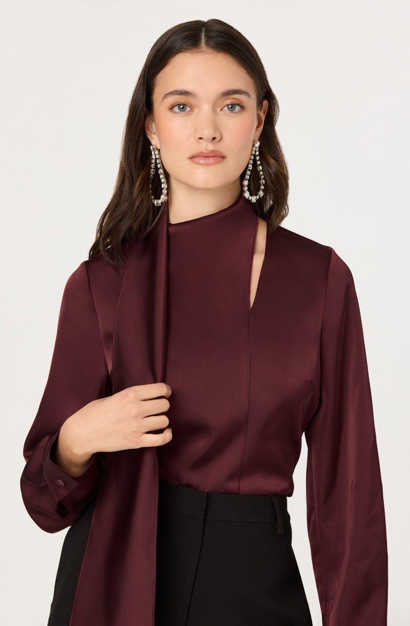 A woman is wearing a long-sleeved, burgundy satin blouse with a high neckline that has a small slit on one side. She is also wearing large, dangling earrings with intricate jeweled details. The blouse appears elegant and has a smooth, shiny texture.