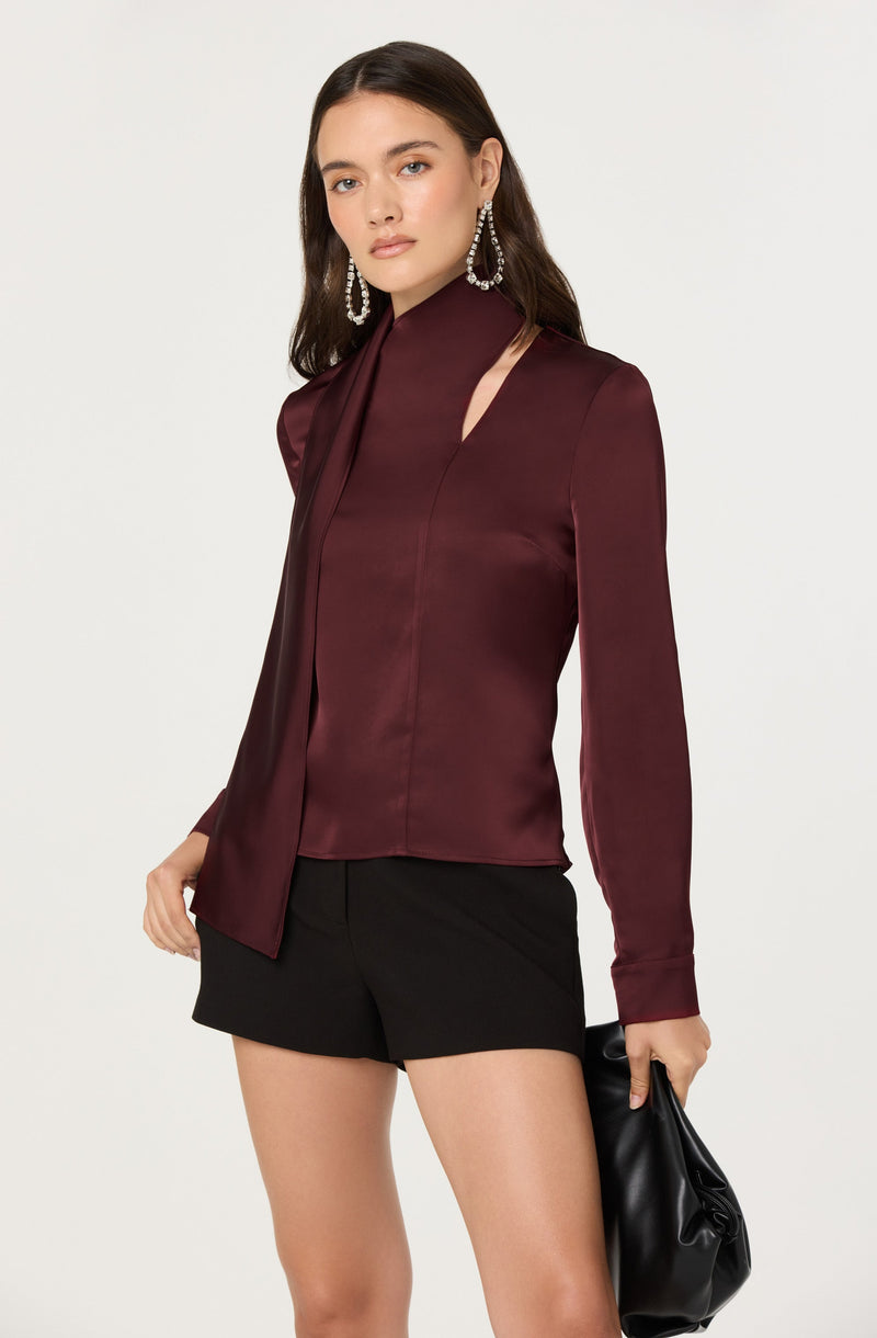 A woman is wearing a long-sleeved, burgundy satin blouse with a high neckline and an asymmetrical slit on the shoulder. She is also wearing black shorts and has large, dangling, chandelier-style earrings. She holds a black clutch bag in her right hand.