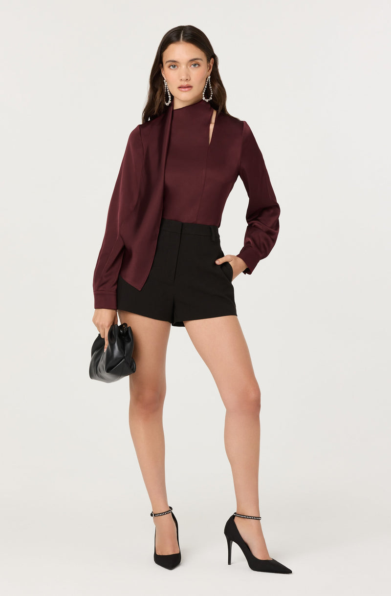 A woman is wearing a long-sleeved, burgundy satin blouse with a asymmetric neckline and a draped detail on one side. She pairs it with high-waisted black shorts and black pointed-toe high heels with ankle straps. She accessorizes with large, dangling earrings and is holding a small, black clutch bag.