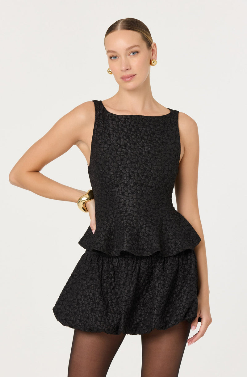 A woman is wearing a sleeveless black textured outfit with a fitted bodice and a peplum detail at the waist, paired with matching textured shorts. She accessorizes with large gold earrings and a gold bracelet on her left wrist.
