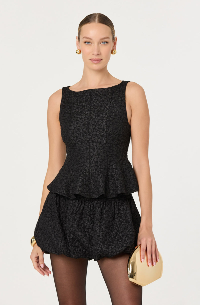 A woman is wearing a sleeveless black textured peplum top with a round neckline and matching textured shorts. She accessorizes with large gold earrings, a gold bracelet, and holds a oval-shaped gold clutch purse. She has her hair styled in a sleek, pulled-back manner.
