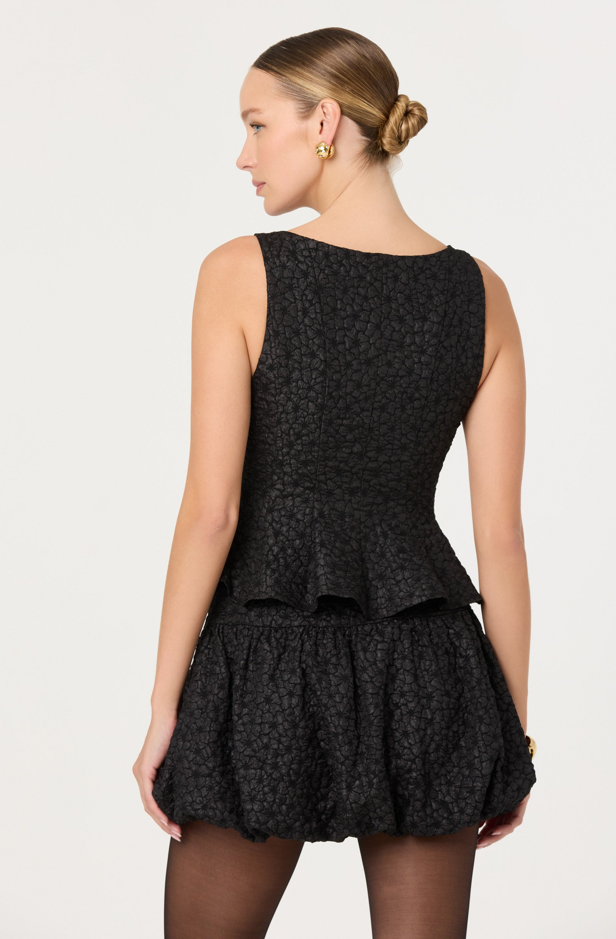 A woman is wearing a sleeveless black top with a textured floral pattern and a peplum hem. She pairs it with matching textured black shorts that have a loose, gathered design.