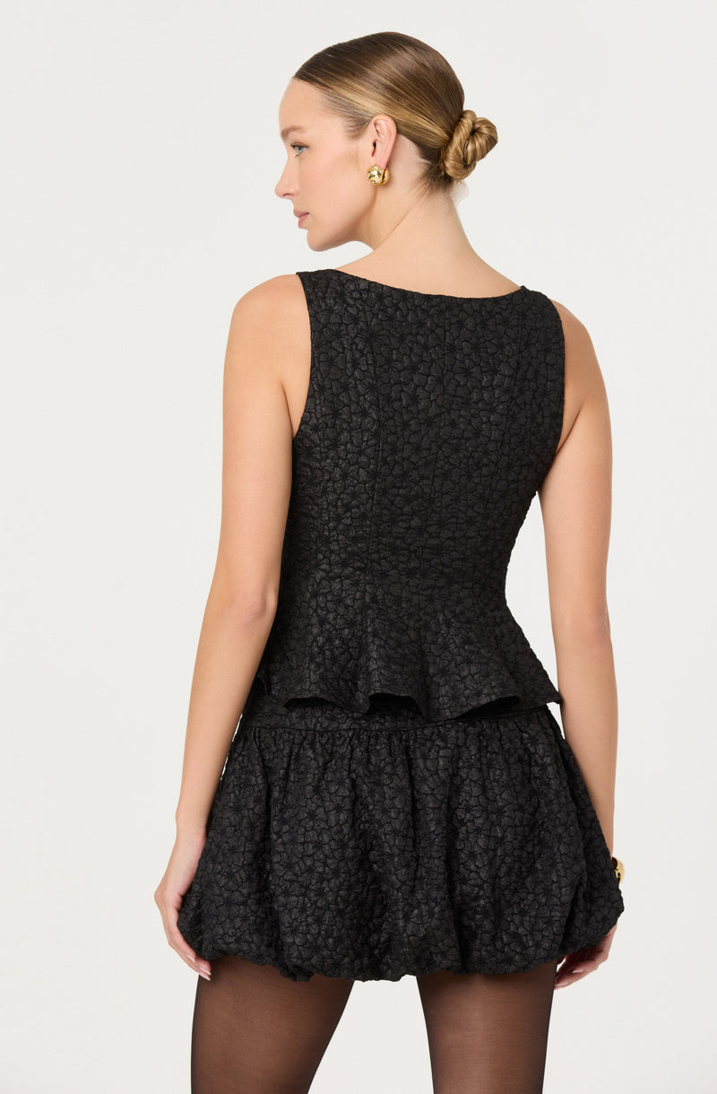 A woman is wearing a sleeveless black top with a textured floral pattern and a peplum hem. She pairs it with matching textured black shorts that have a loose, gathered design.