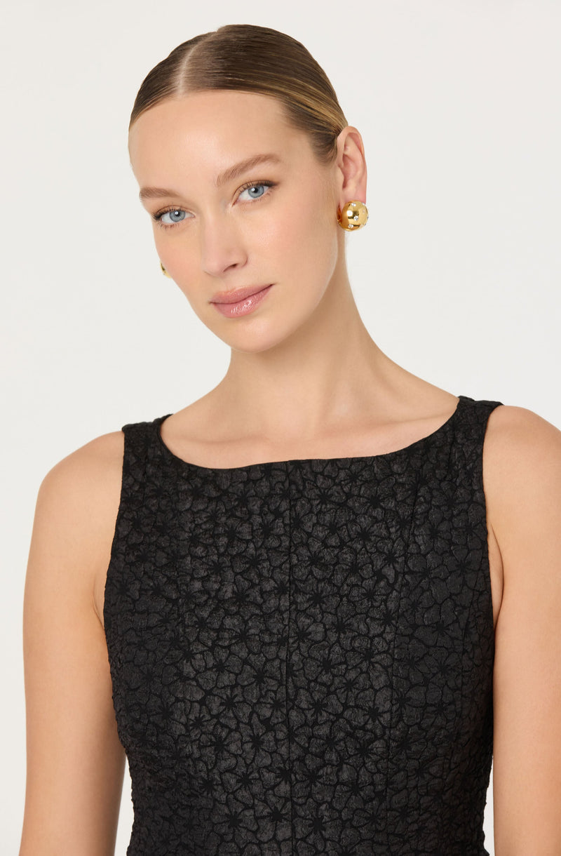 A woman is wearing a sleeveless black dress with a textured, floral pattern. The dress has a round neckline and a fitted silhouette. She is accessorized with large, round, gold earrings.