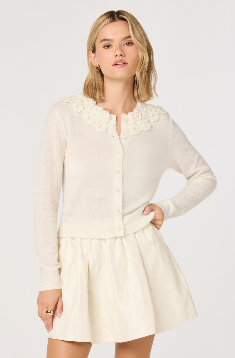 A woman is wearing an ivory or cream-colored cardigan with a floral or textured lace detailing around the neckline. The cardigan has button closures down the front and appears to be made of a soft, knit material. She is also wearing a matching light-colored, possibly satin or silk, knee-length skirt with a gentle sheen.