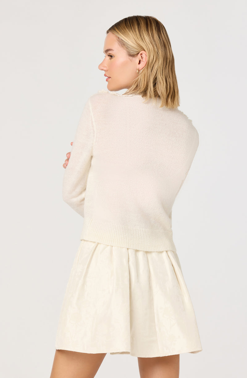 A woman is wearing a soft, light-colored, long-sleeved sweater with a relaxed fit. She pairs it with a matching light-colored, knee-length skirt that has a slight sheen and pleats, creating a classic and elegant look.