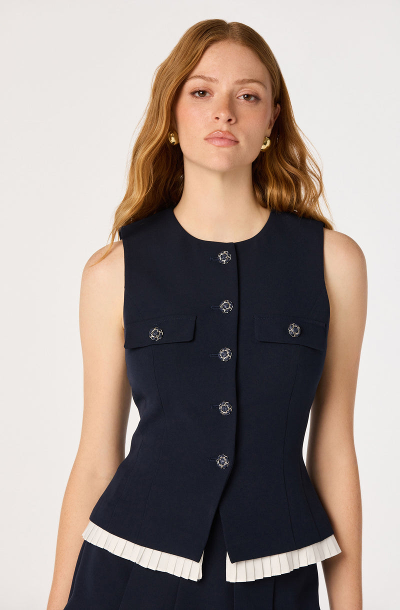 Woman in sleeveless navy vest with decorative buttons, pleated white trim at waist.