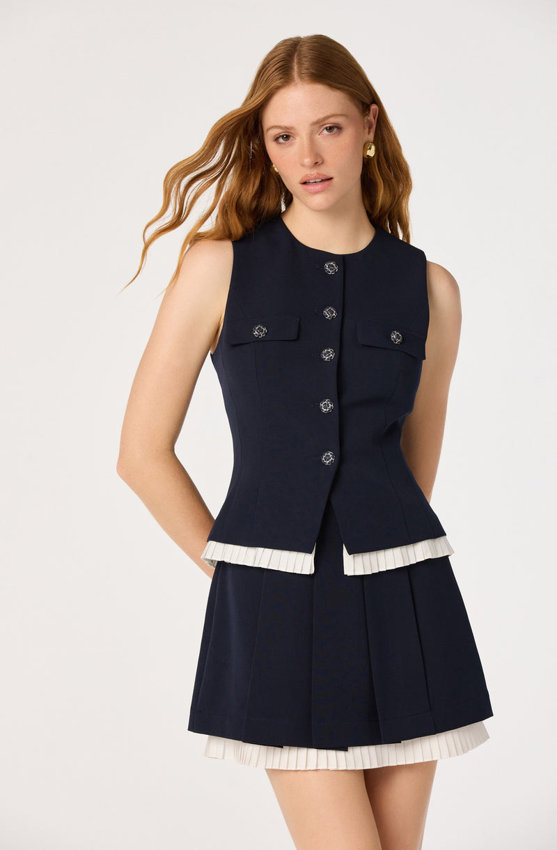Woman wearing a navy sleeveless top with decorative buttons and a pleated navy skirt with white trim.