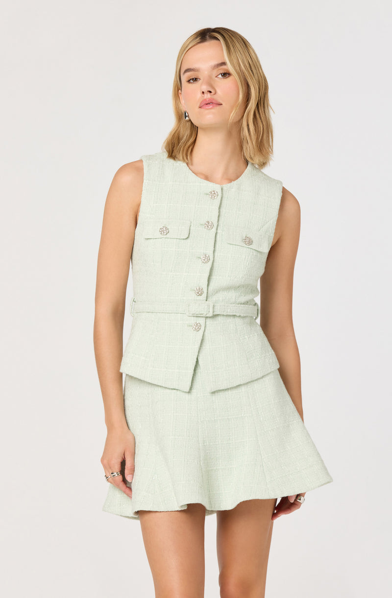 A woman is wearing a sleeveless, pale green, textured two-piece outfit consisting of a cropped vest with decorative buttons and front flap pockets, and a matching skirt with a slight flare.