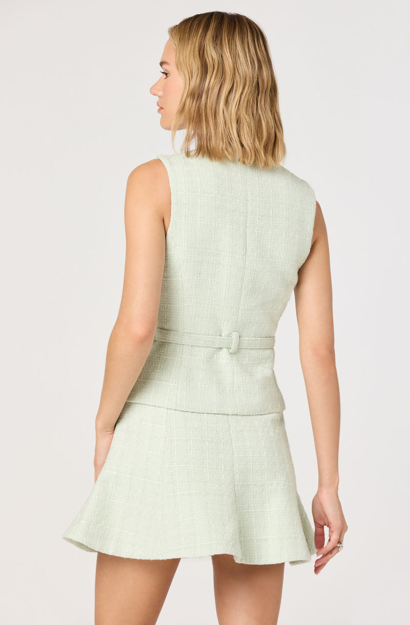 A woman with shoulder-length, wavy blonde hair is wearing a sleeveless, light-colored, textured dress with a fitted bodice and a flared skirt. The dress features a waist detail with a small loop.