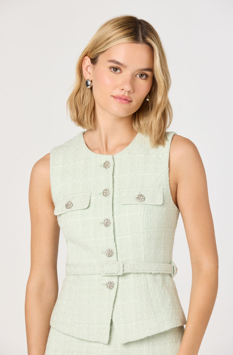 A woman is wearing a sleeveless, light-colored textured vest with decorative buttons and a matching belt at the waist. The vest has two flap pockets on the chest, each secured with a button.