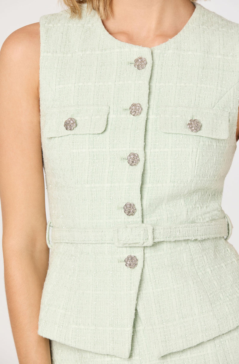 A light green sleeveless dress made of textured fabric, featuring two small flap pockets on the chest, each with a decorative silver button, and matching silver buttons running down the front. The dress also has a matching fabric belt at the waist.