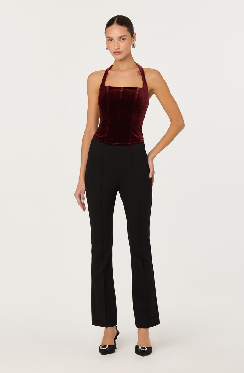 A woman is wearing a sleeveless, fitted burgundy velvet halter top with a square neckline, paired with high-waisted black trousers that have a tailored fit and subtle front seam detailing. She completes her look with black high heels featuring decorative embellishments on the toes.