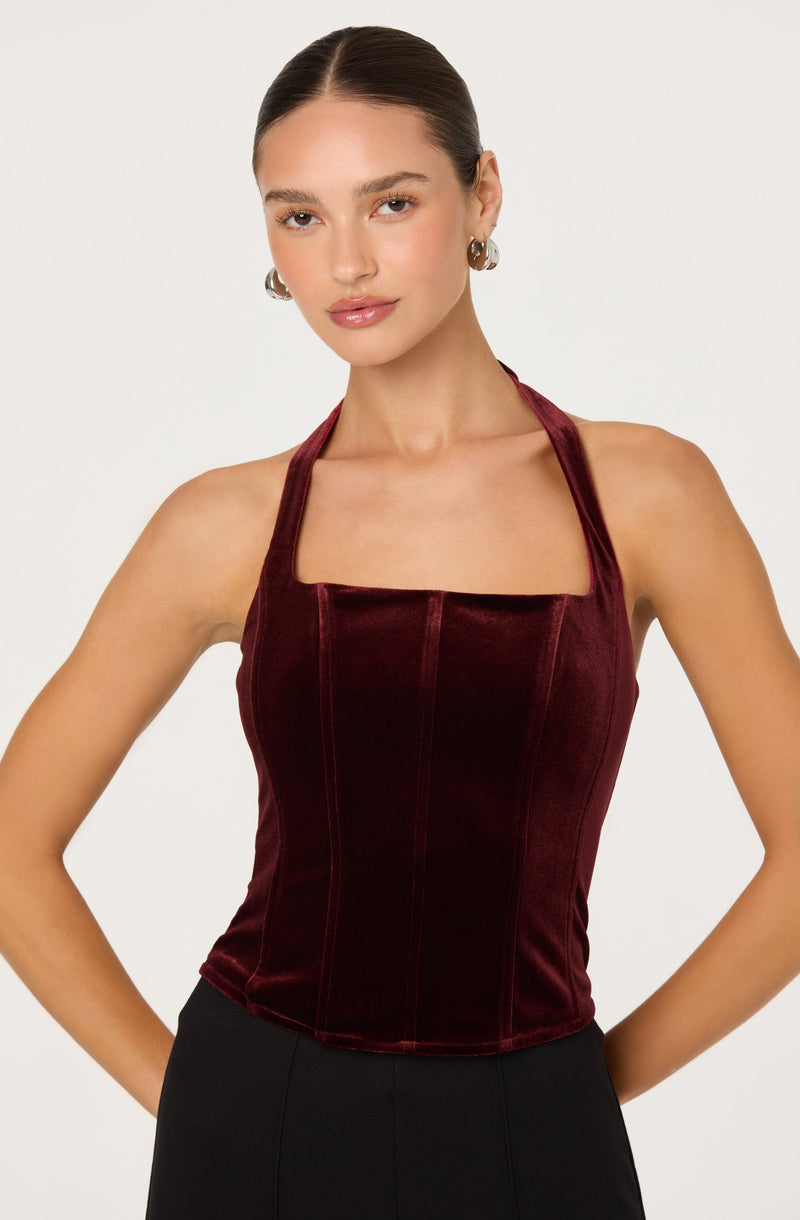 A woman is wearing a sleeveless, fitted, burgundy velvet top with vertical seam details, and a halter neckline. She has dark hair slicked back and is accessorized with large hoop earrings.
