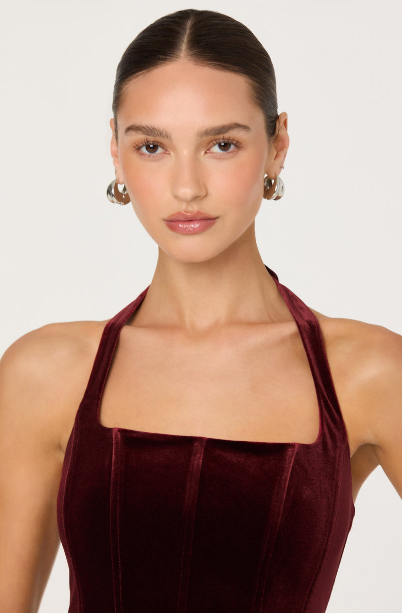 A young woman with fair skin and dark brown hair pulled back, wearing large silver hoop earrings and a dark red velvet dress with a square neckline and wide straps.