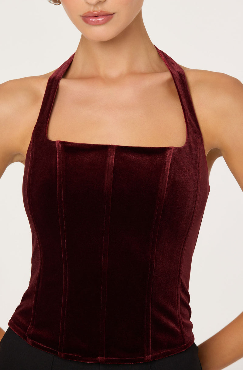 A woman is wearing a fitted, sleeveless halter top made of burgundy velvet fabric. The top features vertical seam details running down the front, creating a structured and tailored appearance. The neckline is square-shaped, and the wide halter straps go around her neck, leaving her shoulders and arms bare.