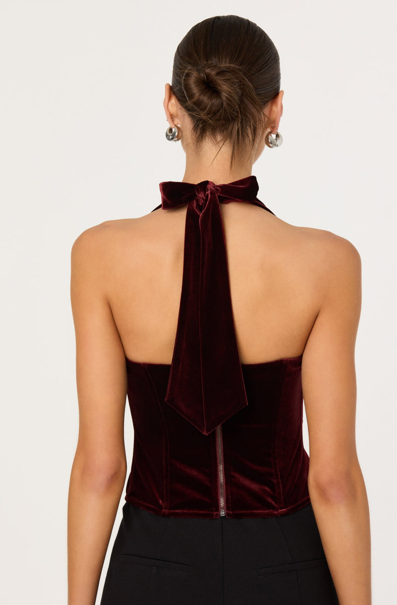 A woman is wearing a strapless, deep burgundy velvet top with a large bow tied around her neck, which extends down her back. The top has a zipper closure at the back. She accessorizes with large, circular silver earrings and has her hair styled in a neat bun.