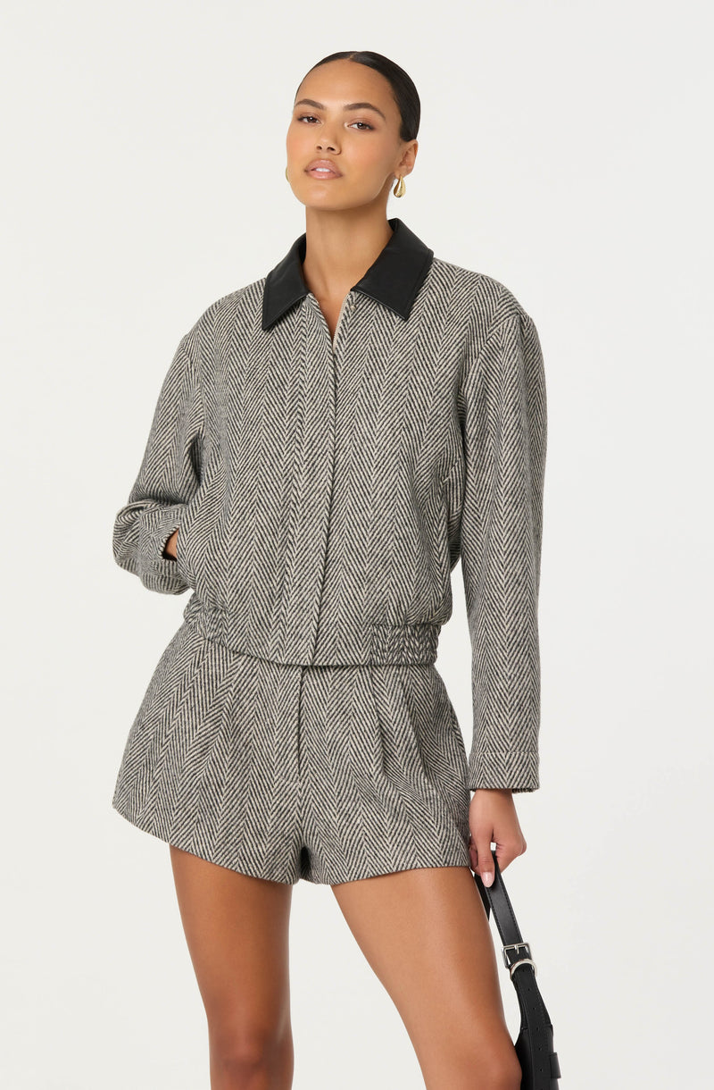 A woman is wearing a matching blazer and shorts set in a herringbone pattern with a black collar on the blazer. The blazer has a relaxed fit with long sleeves, and the shorts are high-waisted with an elastic waistband. The fabric appears to be woven in a classic tweed-like pattern with neutral tones.
